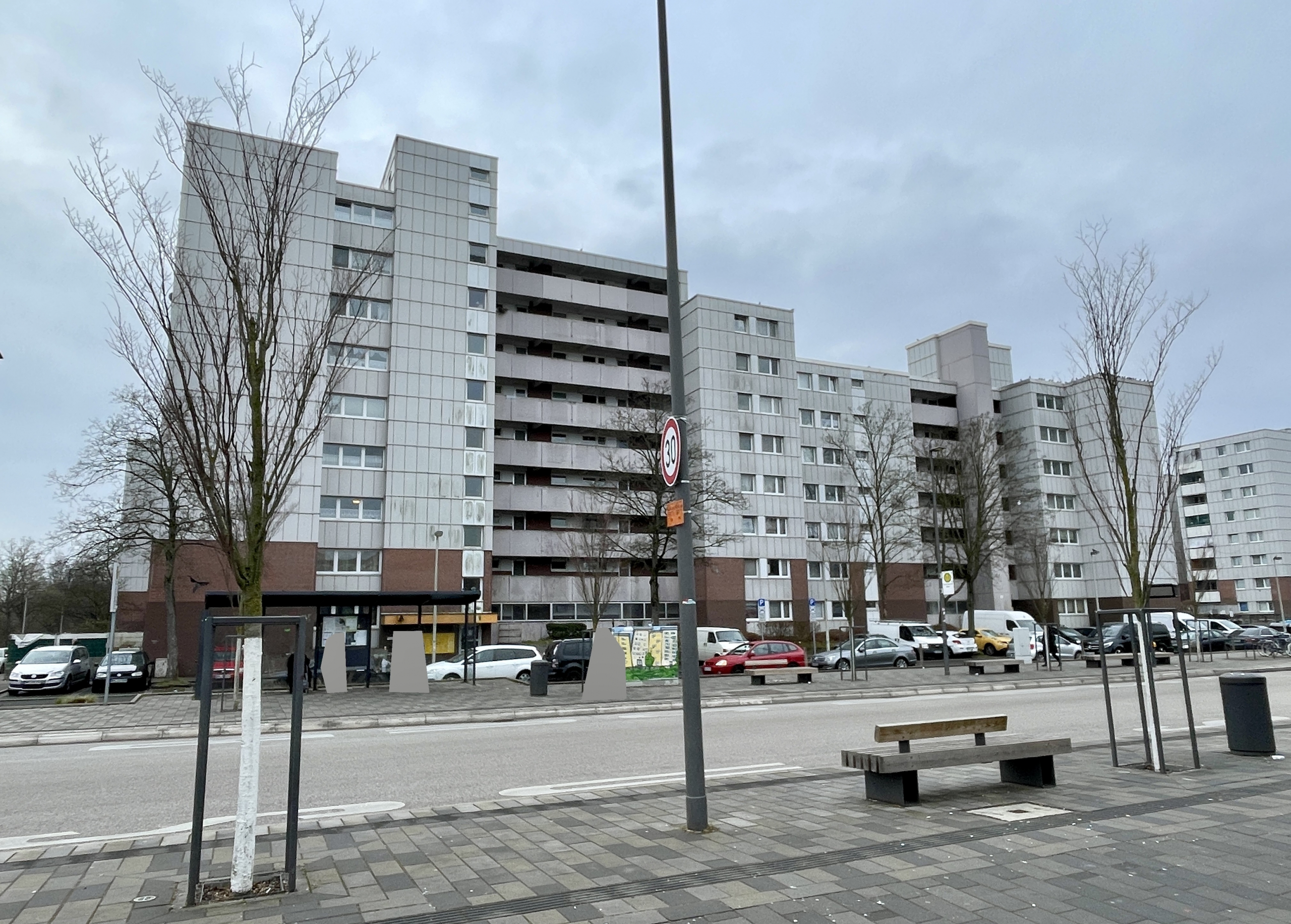 Foto des Wohngebäudes an der Oppelner Straße 51 in Bonn Tannenbusch. Im Vordergrund zwei Bäume, eine Laterne, eine Bank und ein Mülleimer. Im Hintergrund das neunstöckige Gebäude.