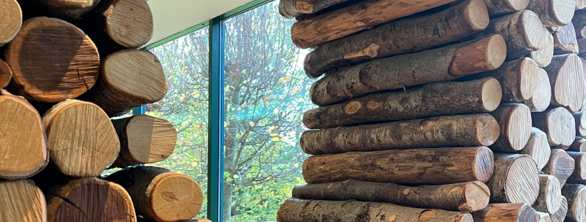 Foto von Rundhölzern, die so gestapelt sind, dass sie eine runde Öffnung freilassen. Durch die Öffnung sieht man in den Raum dahinter und dort durch ein Fenster ins Grüne.